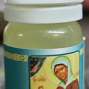 May include: A small, clear glass bottle with a white cap. The bottle contains a yellow liquid and has a label with a blue background and a picture of a woman in a white head covering. The label also has the text "Святой России" in Russian.