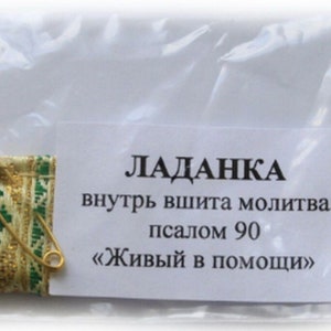 May include: A small, green and gold fabric pouch with a safety pin attached. The pouch is labeled "Ладанка" which translates to "amulet" in English. The label also says "внутрь вышита молитва псалом 90 "Живый в помощи" which translates to "Inside is embroidered a prayer, Psalm 90 "Living in help."