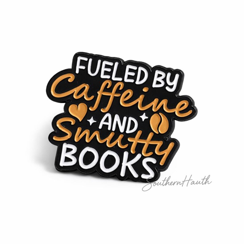 May include: A black enamel pin with gold and white lettering that reads FUELED BY Caffeine AND Smutty BOOKS. The design includes a small gold heart and a coffee bean icon, with the text arranged in a stacked, stylized layout on a black background.