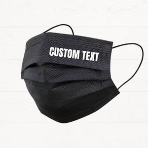 Puede incluir: Mascarilla negra con presillas para las orejas. La mascarilla tiene pliegues y las palabras "CUSTOM TEXT" impresas en blanco en la parte delantera. El fondo es un grano de madera blanco.