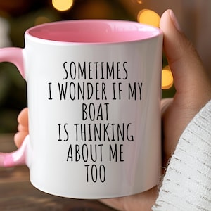 May include: White ceramic mug with a pink interior and handle. The mug features the text "SOMETIMES I WONDER IF MY BOAT IS THINKING ABOUT ME TOO" in black lettering. The mug is held by a person.