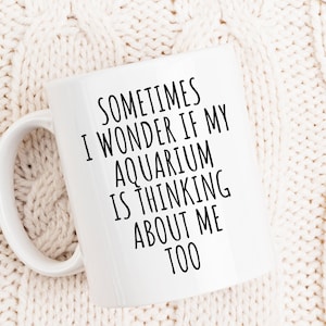 May include: A white ceramic mug with a curved handle. The mug features black text that reads, "SOMETIMES I WONDER IF MY AQUARIUM IS THINKING ABOUT ME TOO." The mug is set against a cream-colored knitted background.