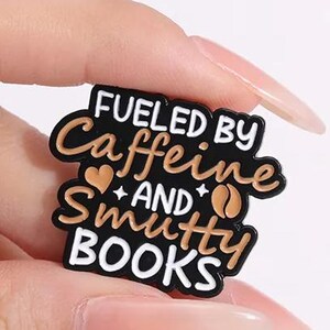 May include: A black enamel pin with white and tan lettering that reads FUELED BY Caffeine AND Smutty BOOKS. The design includes a small tan heart and a tan coffee bean icon, all outlined in black metal with a smooth, glossy finish.