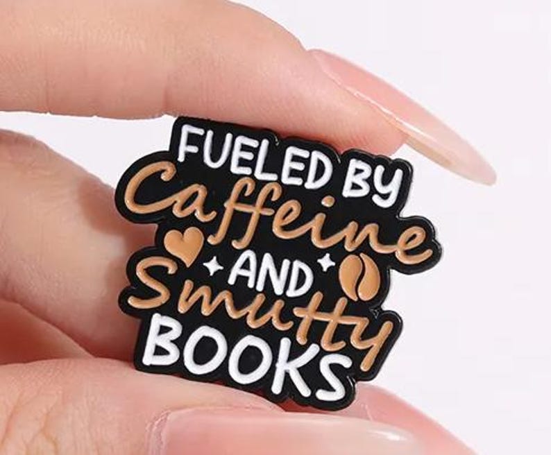 May include: A black enamel pin with white and tan lettering that reads FUELED BY Caffeine AND Smutty BOOKS. The design includes a small tan heart and a tan coffee bean icon, all outlined in black metal with a smooth, glossy finish.