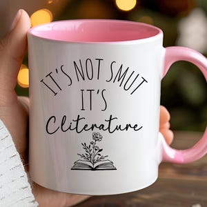 Op de afbeelding: Witte keramische mok met roze binnenkant en oor. De mok heeft de zwarte tekst "IT'S NOT SMUT IT'S Cliterature" met een bloemenillustratie. Een hand houdt de mok vast.