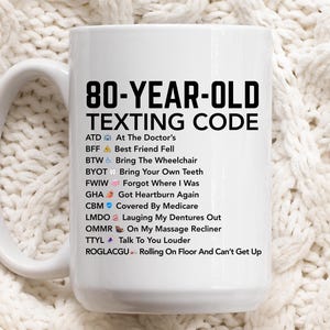 May include: White ceramic mug with the text "80-YEAR-OLD TEXTING CODE" in black. The mug lists common texting abbreviations with their meanings, such as "ATD At The Doctor's" and "BFF Best Friend Fell". The mug has a white handle.