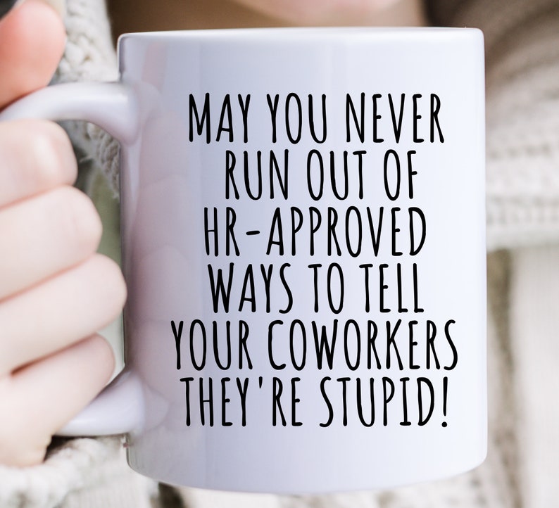 May include: White ceramic mug with a black humorous quote: "MAY YOU NEVER RUN OUT OF HR-APPROVED WAYS TO TELL YOUR COWORKERS THEY'RE STUPID!" The mug has a handle and is held by a person.