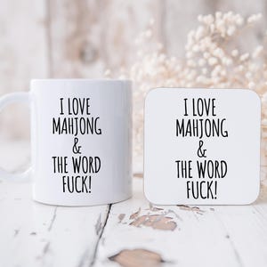 May include: White ceramic mug and square coaster with the text "I LOVE MAHJONG & THE WORD FUCK!" in black. The mug has a handle and the coaster is square. The items are on a white, distressed wooden surface.