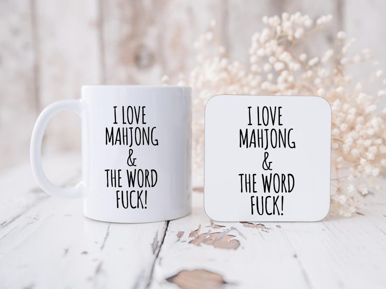 May include: White ceramic mug and square coaster with the text "I LOVE MAHJONG & THE WORD FUCK!" in black. The mug has a handle and the coaster is square. The items are on a white, distressed wooden surface.