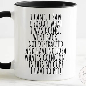 May include: A white ceramic mug with a black handle and the text "But First, Coffee" in a bold, black font. The mug is filled with a dark liquid, likely coffee, and sits on a light-colored surface.