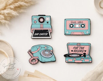 May include: Four enamel pins featuring retro designs in pastel blue and pink. One is a camera with the text SAY CHEESE, one is a cassette tape with GOOD VIBES, one is a rotary telephone, and one is a record player with FOR THE RECORD.