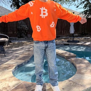 May include: An orange sweatshirt with multiple white Bitcoin symbols. The long-sleeved crew neck shirt is worn with light blue jeans and white sneakers. The person is standing in front of a pool.