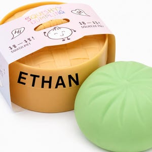 May include: A light green round squishy dumpling toy next to a yellow plastic steamer basket container. The container features a white paper sleeve printed with the text SQUISHY DUMPLING, HI, 18-11!, SQUEEZE ME!, and the name ETHAN in bold black letters.