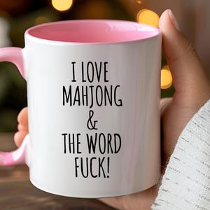 May include: White ceramic mug with a pink interior and handle. The mug features the text "I LOVE MAHJONG & THE WORD FUCK!" in black, bold letters. The mug is held by a person.