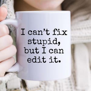 Peut inclure: Mug en céramique blanche avec le texte "I can't fix stupid, but I can edit it." en noir, police sans-serif. La tasse est tenue par une personne portant un pull blanc. La tasse a une anse standard.