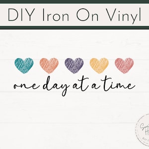May include: DIY iron-on vinyl design with five coloured hearts: teal, orange, purple, yellow, and pink. The phrase "one day at a time" is written in a cursive font below the hearts. A circular logo with text is in the bottom right corner.