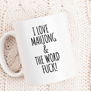 May include: White ceramic mug with the text "I LOVE MAHJONG & THE WORD FUCK!" in black. The mug has a curved handle and is set against a cream-colored knitted background.