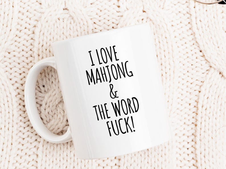 May include: White ceramic mug with the text "I LOVE MAHJONG & THE WORD FUCK!" in black. The mug has a curved handle and is set against a cream-colored knitted background.