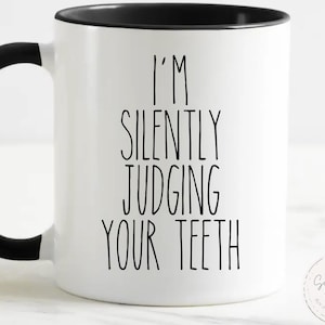 Puede incluir: Taza de cerámica blanca con asa y borde negros. La taza presenta el texto "I'M SILENTLY JUDGING YOUR TEETH" en letras negras y en negrita. Un pequeño logotipo circular es visible en el lado derecho de la taza.