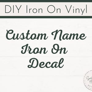 May include: DIY iron-on vinyl decal with the text "Custom Name Iron On Decal" in a dark green script font. The text is displayed on a white background with a wood grain pattern. A small circular logo is in the bottom right corner.
