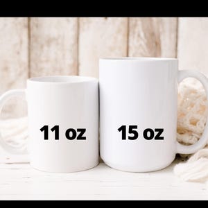 May include: Two white ceramic mugs, one labeled "11 oz" and the other "15 oz". The mugs have a simple, clean design with a curved handle. The background is a light wooden surface.