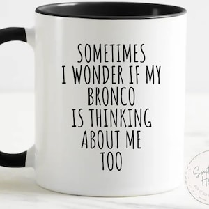 May include: White ceramic mug with a black handle and rim. The mug features the text "SOMETIMES I WONDER IF MY BRONCO IS THINKING ABOUT ME TOO" in black. A small circular logo is on the right side.