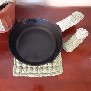 Könnte beinhalten: Eine schwarze gusseiserne Bratpfanne mit zwei passenden hellgrünen gehäkelten Griffschützern und einem quadratischen Topflappen. Die Pfanne steht auf dem Topflappen. Die Griffschützer sind auf den Pfannengriffen platziert. Die Artikel befinden sich auf einer dunklen Holzoberfläche.