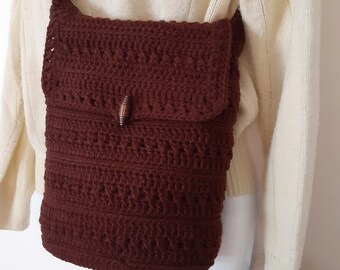 Könnte beinhalten: Eine braune gehäkelte Umhängetasche mit einem Holz-Knopfverschluss. Die Tasche wird über einem weißen Pullover getragen.