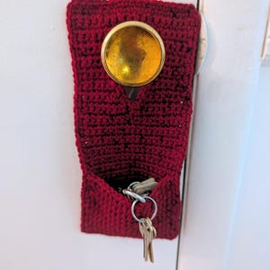 May include: A dark red crocheted key holder featuring a circular cutout at the top that fits over a round gold-coloured door knob. The rectangular fabric pouch has an open pocket at the bottom containing a metal key ring with several silver keys.