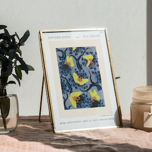 Könnte beinhalten: Ein goldfarbener Bilderrahmen mit einem blauen und gelben Blumendruck. Das Bild steht auf einem Tisch mit einer Vase mit grünen Blättern und einer Kerze.