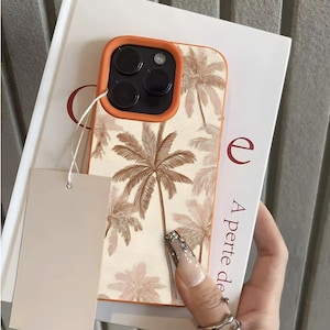 Puede incluir: Funda de teléfono naranja y crema con diseño de palmeras. La funda tiene un borde elevado alrededor de la cámara y una etiqueta en blanco adjunta. La funda del teléfono está sobre un libro con el título "A perte de".