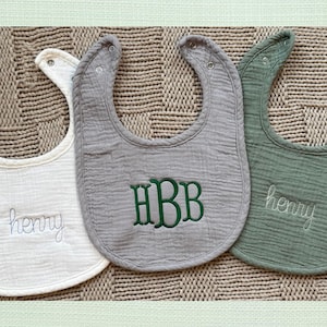May include: Three baby bibs in white, gray, and green, each with a unique embroidered design. The white bib has the name "henry" embroidered in a cursive font. The gray bib features the initials "HBB" in a block font. The green bib also has the name "henry".