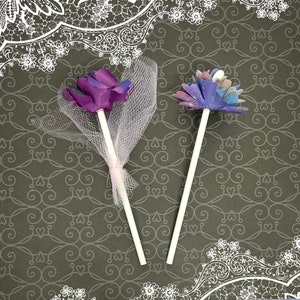 Puede incluir: Dos adornos para pasteles con forma de flor. Uno es morado con un lazo de tul blanco y el otro es azul y morado con un dise&ntilde;o de galaxia.