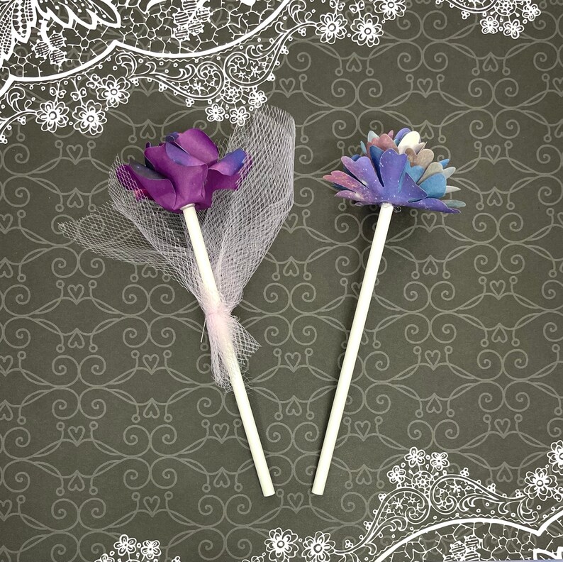 Puede incluir: Dos adornos para pasteles con forma de flor. Uno es morado con un lazo de tul blanco y el otro es azul y morado con un dise&ntilde;o de galaxia.