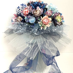 May include: A bouquet of colorful paper flowers with wrapped lollipops in the center. The bouquet is wrapped in a white and blue tulle with a blue ribbon with butterfly designs.