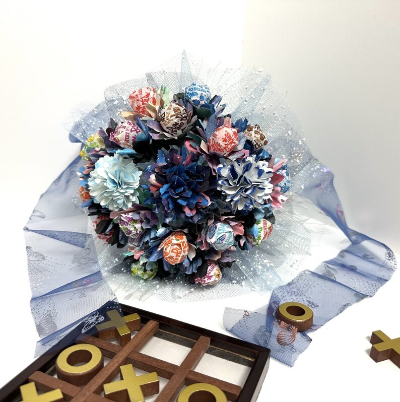 May include: A bouquet of blue, pink, and white paper flowers with candy wrapped in colorful paper. The bouquet is wrapped in blue tulle and sits on a white surface.