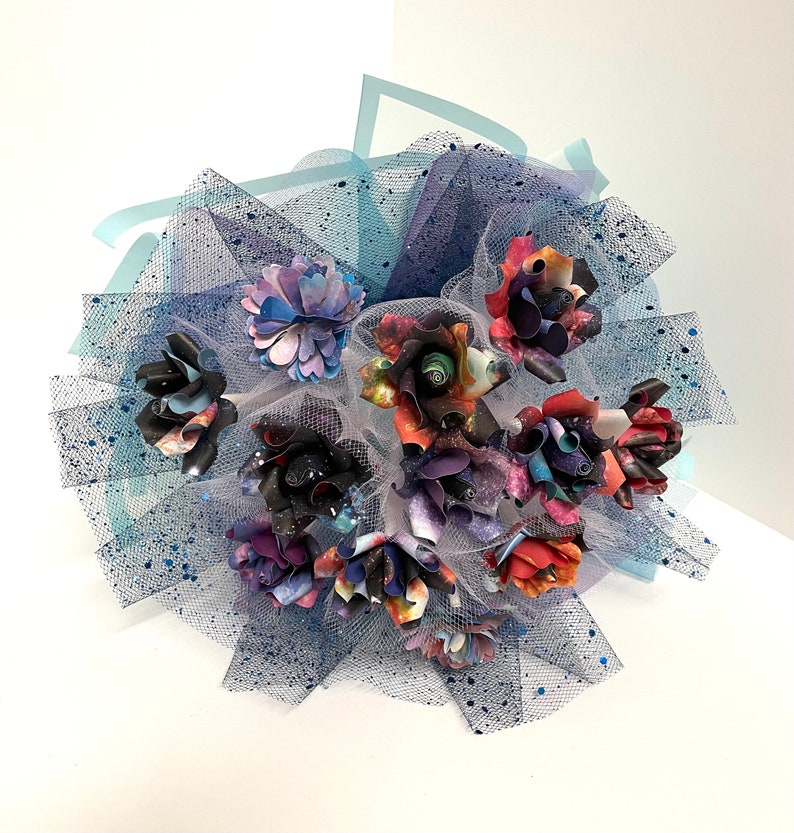 Puede incluir: Un ramo de flores de papel con estampado de galaxia, arreglado en un tul azul con brillantina azul.