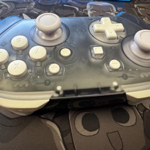 May include: A transparent gray video game controller with white buttons and joysticks. The controller has a cross-shaped directional pad, four face buttons, and two analog sticks. The buttons are labeled with letters and symbols.