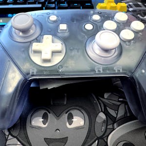May include: A transparent blue video game controller with white buttons and gray joysticks. The controller has a D-pad, face buttons, and shoulder buttons. The controller is resting on a black and white graphic of a cartoon character.