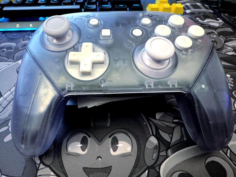 May include: A transparent blue video game controller with white buttons and gray joysticks. The controller has a D-pad, face buttons, and shoulder buttons. The controller is resting on a black and white graphic of a cartoon character.