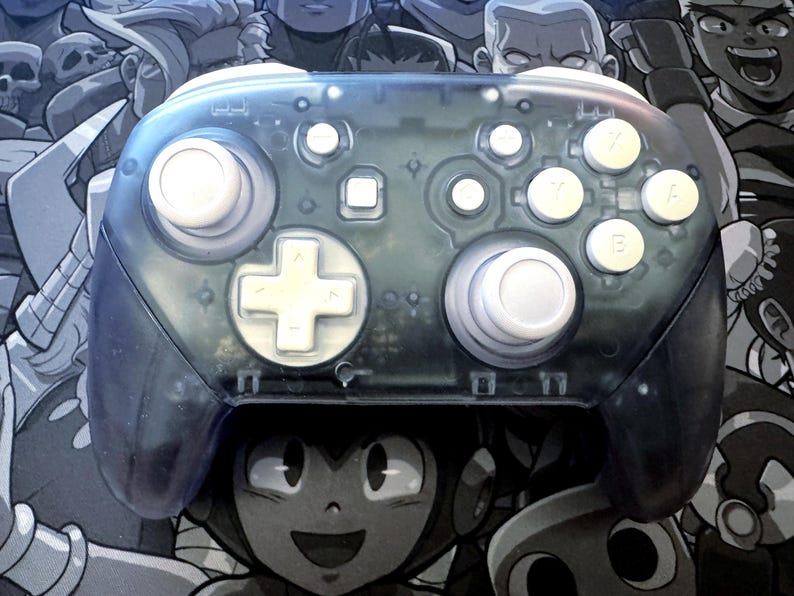 May include: A transparent blue video game controller with white buttons and a white directional pad. The controller has a clear shell, revealing the internal components. The background features a grayscale illustration of video game characters.