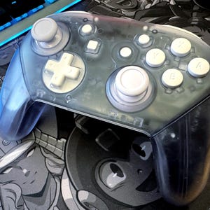 May include: A transparent blue video game controller with white buttons and joysticks. The controller has a D-pad, face buttons, and shoulder buttons. The controller is on a gaming mousepad with a cartoon design.