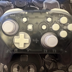 May include: A transparent gray video game controller with white buttons and a white D-pad. The controller has a clear shell, allowing a view of the internal components. The buttons are labeled with letters and symbols.
