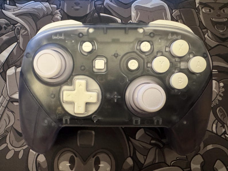 May include: A transparent gray video game controller with white buttons and a white D-pad. The controller has a clear shell, allowing a view of the internal components. The buttons are labeled with letters and symbols.