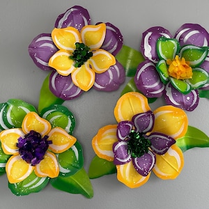 May include: Three colorful, handcrafted flower decorations. Each flower features a vibrant mix of yellow, purple, and green petals with a textured center. The flowers are accompanied by green leaves, creating a cheerful, decorative arrangement.