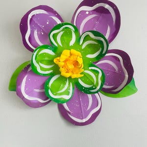 May include: A purple paper flower with white and green accents. The flower has a yellow center and is decorated with white dots.