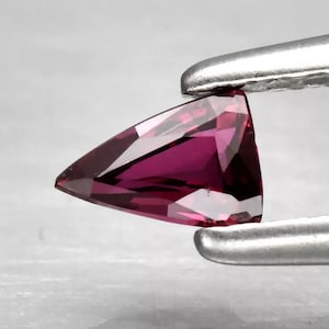 Op de afbeelding: Een gefacetteerde, driehoekige edelsteen in tinten diep magenta en paars wordt vastgehouden door een zilveren pincet. De edelsteen heeft scherpe hoeken en een gepolijst oppervlak, dat licht reflecteert. De edelsteen is een levendig, opvallend stuk.