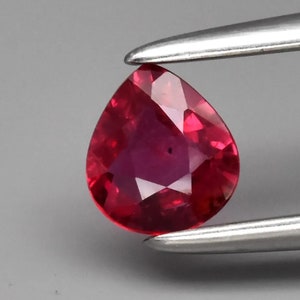 Peut inclure: Une pierre précieuse de rubis de forme poire avec une couleur rouge foncé. La pierre précieuse est tenue dans une pince.