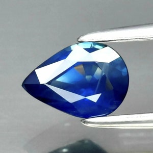 May include: A pear-shaped blue sapphire gemstone with a faceted cut. The gemstone is held by a pair of tweezers.