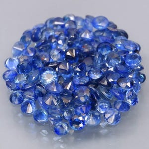 May include: A pile of round, blue gemstones with multiple facets. The gems are in varying shades of blue, from pale to deep, and reflect light. The arrangement is clustered, creating a sparkling display. These gems are likely for jewellery or decorative use.
